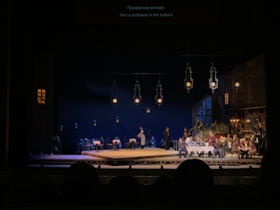 A stage performance with a group of people, some seated at tables with white tablecloths and others standing. The set is designed to look like an outdoor night scene with several hanging lantern-style lights and a dark background. The audience is visible in the foreground, watching the performance.