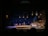 A stage performance with a group of people, some seated at tables with white tablecloths and others standing. The set is designed to look like an outdoor night scene with several hanging lantern-style lights and a dark background. The audience is visible in the foreground, watching the performance.