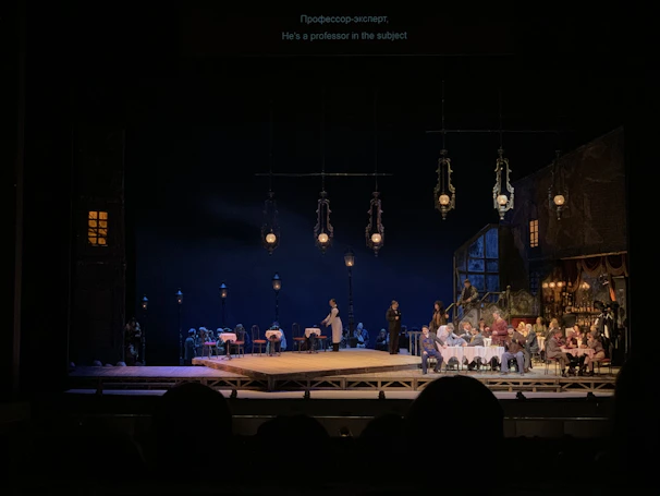 Evening outdoor theatrical performance with golden stage lights and a captivated rural audience.