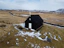 A small black wooden church stands alone in a snow-dusted, barren landscape. The ground is mostly covered with patches of snow, and a winding road leads from the distance to the church, emphasizing its isolation. Surrounding hills are mostly bare with a distant view of mountains under a partly cloudy sky.