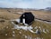 A small black wooden church stands alone in a snow-dusted, barren landscape. The ground is mostly covered with patches of snow, and a winding road leads from the distance to the church, emphasizing its isolation. Surrounding hills are mostly bare with a distant view of mountains under a partly cloudy sky.