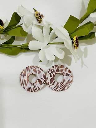 Two round, flat discs with a textured pattern resembling animal print or bark lie against a white background. Above them are artificial white flowers with green leaves. The flowers have brown and yellow centers, and the arrangement appears minimalistic and decorative.