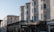 A street scene featuring a double-decker bus marked 'Brighton Station' passing by several multi-story residential buildings with bay windows. The ground level has various storefronts, one with a 'To Let' sign. The architecture suggests a vintage style with neutral colors.
