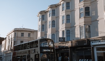 A street scene featuring a double-decker bus marked 'Brighton Station' passing by several multi-story residential buildings with bay windows. The ground level has various storefronts, one with a 'To Let' sign. The architecture suggests a vintage style with neutral colors.