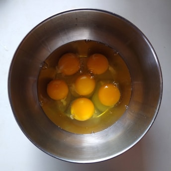 A metal bowl contains six raw egg yolks with the whites visible, placed on a light-colored surface.
