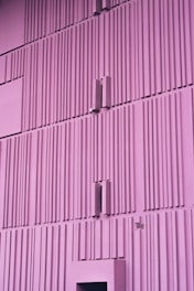 A minimalistic, vertically-aligned architecture with a purple hue. The facade features a repetitive pattern of rectangular ridges and protrusions, giving it a modern, geometric appearance.