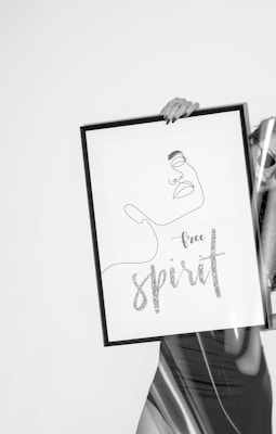 A minimalist, black and white line drawing of a face with the words 'free spirit' in cursive below it. The drawing is held in a black frame by a hand with painted nails. Part of a person wearing a sleek dress is visible to the side, partially obscured by the frame.