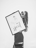 A black and white image featuring a person holding a large framed artwork. The artwork contains an abstract line drawing of a face accompanied by the words 'Free Spirit'. The figure appears stylish, wearing sunglasses with a calm demeanor.