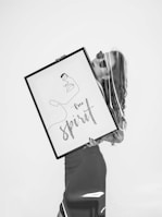 A black and white image featuring a person holding a large framed artwork. The artwork contains an abstract line drawing of a face accompanied by the words 'Free Spirit'. The figure appears stylish, wearing sunglasses with a calm demeanor.