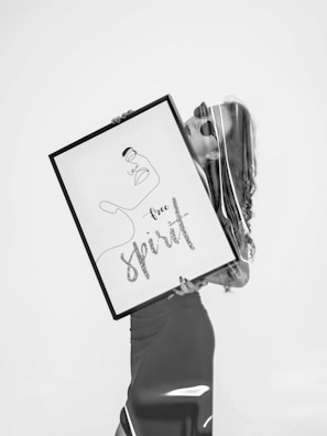 A black and white image featuring a person holding a large framed artwork. The artwork contains an abstract line drawing of a face accompanied by the words 'Free Spirit'. The figure appears stylish, wearing sunglasses with a calm demeanor.