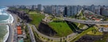 A coastal cityscape featuring high-rise buildings lining the shore, with a green cliffside park dividing the urban area from the ocean. Roads curve along the coast, and a bridge spans the green space. The ocean is to the left with waves crashing against the shore.