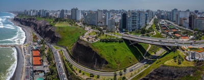 A coastal cityscape featuring high-rise buildings lining the shore, with a green cliffside park dividing the urban area from the ocean. Roads curve along the coast, and a bridge spans the green space. The ocean is to the left with waves crashing against the shore.