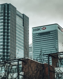 Two tall skyscrapers with glass facades dominate the skyline. Each building prominently displays the logos of financial institutions. In the foreground, there are modern residential buildings with brick walls and metal frameworks. The sky is overcast with gray clouds.
