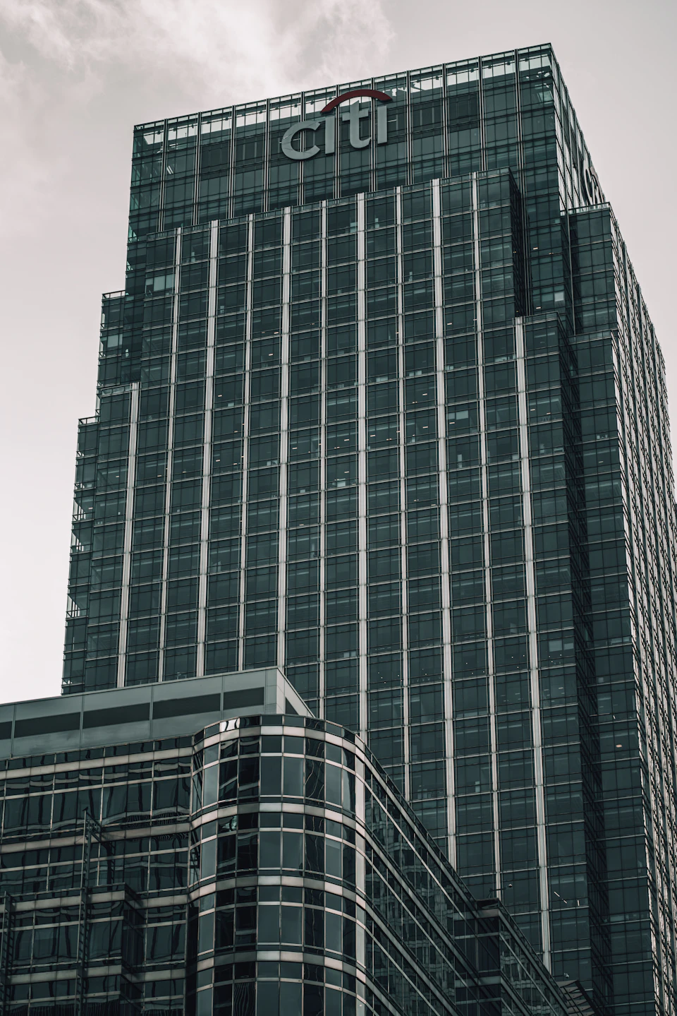 The image shows a tall, modern glass and steel building with a curved facade that reflects the surrounding environment. The building is adorned with numerous windows, adding to its sleek and contemporary design. Atop the structure, there's a large sign that reads "CITI", indicating the presence of a financial institution within the premises.