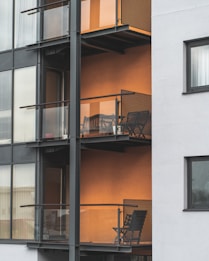 The image depicts a modern apartment building featuring multiple balconies with minimalist metal railings and glass panels. Each balcony contains a small outdoor seating area consisting of a chair and a table, set against an orange-colored wall that adds warmth to the exterior. The building's facade combines both glass and concrete elements, providing a sleek urban appearance.