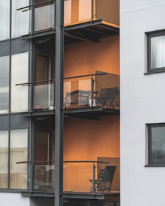 The image depicts a modern apartment building featuring multiple balconies with minimalist metal railings and glass panels. Each balcony contains a small outdoor seating area consisting of a chair and a table, set against an orange-colored wall that adds warmth to the exterior. The building's facade combines both glass and concrete elements, providing a sleek urban appearance.