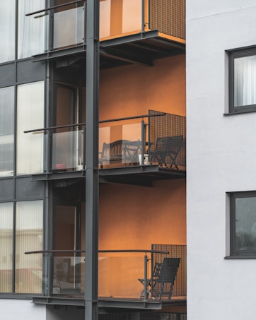 The image depicts a modern apartment building featuring multiple balconies with minimalist metal railings and glass panels. Each balcony contains a small outdoor seating area consisting of a chair and a table, set against an orange-colored wall that adds warmth to the exterior. The building's facade combines both glass and concrete elements, providing a sleek urban appearance.