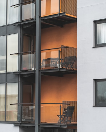 The image depicts a modern apartment building featuring multiple balconies with minimalist metal railings and glass panels. Each balcony contains a small outdoor seating area consisting of a chair and a table, set against an orange-colored wall that adds warmth to the exterior. The building's facade combines both glass and concrete elements, providing a sleek urban appearance.