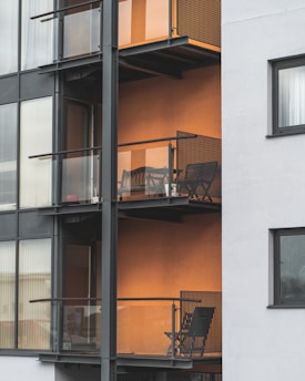 The image depicts a modern apartment building featuring multiple balconies with minimalist metal railings and glass panels. Each balcony contains a small outdoor seating area consisting of a chair and a table, set against an orange-colored wall that adds warmth to the exterior. The building's facade combines both glass and concrete elements, providing a sleek urban appearance.