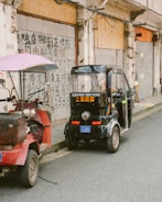 Two small vehicles are parked beside a wall with Chinese writing. The first vehicle is a red scooter-like vehicle with a purple canopy, while the second is a small black electric tricycle with its doors open. The wall in the background is aged and shows visible electrical wires.