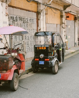 Two small vehicles are parked beside a wall with Chinese writing. The first vehicle is a red scooter-like vehicle with a purple canopy, while the second is a small black electric tricycle with its doors open. The wall in the background is aged and shows visible electrical wires.