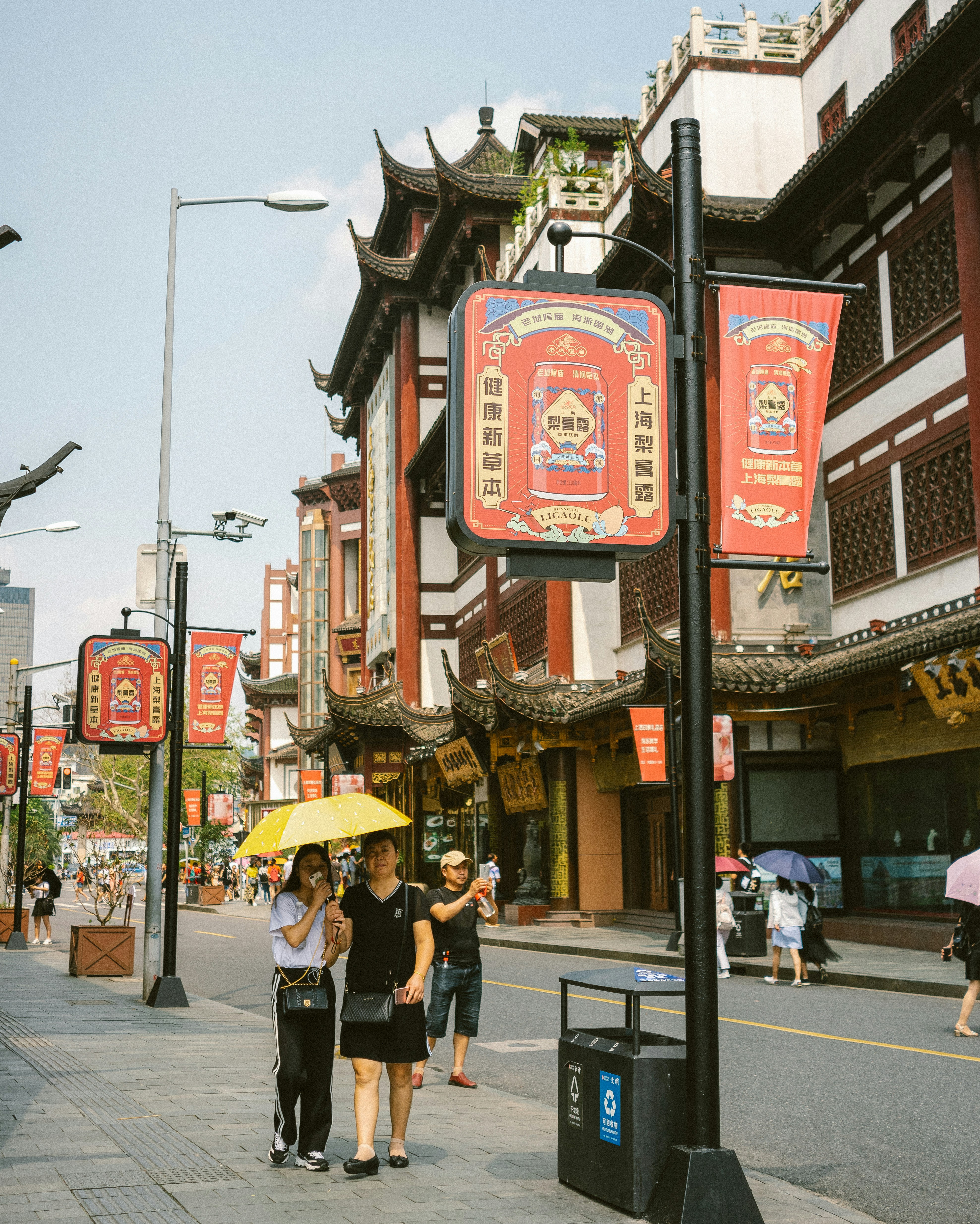 Photograph of a Shanghai street lined with ornate façades and red lantern banners, with pedestrians and a bright yellow umbrella in the foreground.