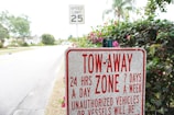 A tow-away zone sign prominently displays information about towing policies 24 hours a day, 7 days a week, against unauthorized vehicles or vessels. Behind the sign, there is a speed limit sign indicating 25 mph and a road lined with trees and shrubs.