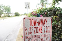 A tow-away zone sign prominently displays information about towing policies 24 hours a day, 7 days a week, against unauthorized vehicles or vessels. Behind the sign, there is a speed limit sign indicating 25 mph and a road lined with trees and shrubs.
