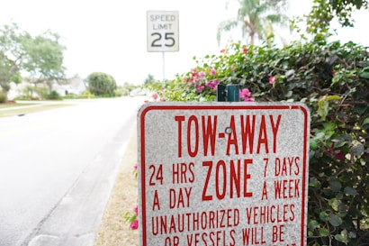 A tow-away zone sign prominently displays information about towing policies 24 hours a day, 7 days a week, against unauthorized vehicles or vessels. Behind the sign, there is a speed limit sign indicating 25 mph and a road lined with trees and shrubs.