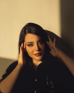 A woman with long dark hair gazes at the camera with a serene expression. Her hands are gently placed near her face and golden sunlight softly illuminates her features, creating a play of light and shadow.