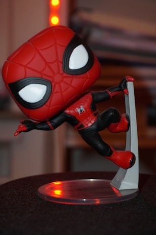 A stylized vinyl figure of a superhero character wearing a red and black suit with a web pattern on the mask. The figure is posed dynamically, balancing on one leg with an arm extended. Its large eyes are outlined in black, giving it a cartoonish appearance. The background is blurred and features hints of warmly colored lights.