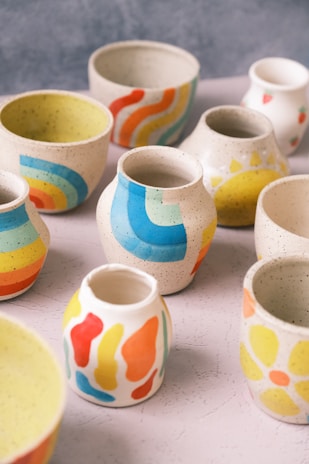 Various ceramic pots and vessels are displayed, each adorned with colorful, abstract patterns and designs, including stripes, waves, and floral motifs. The pots are placed on a textured surface, and their vibrant colors contrast with the neutral tones of the pottery itself.