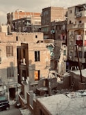 An urban landscape featuring old, weathered buildings in a state of disrepair. Some structures have partially collapsed walls, and balconies and windows are in various stages of decay. A variety of textures and materials such as brick, stone, and concrete are visible. Laundry hangs from several windows, and satellite dishes and cables are visible on rooftops. The scene suggests an area that has experienced neglect or damage.