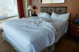 A large, unmade bed with white sheets and multiple pillows lies in a room with a wooden headboard and side tables. A table lamp can be seen on one of the side tables, and a window with a cityscape view is partially covered by red curtains.
