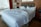A large, unmade bed with white sheets and multiple pillows lies in a room with a wooden headboard and side tables. A table lamp can be seen on one of the side tables, and a window with a cityscape view is partially covered by red curtains.