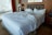A large, unmade bed with white sheets and multiple pillows lies in a room with a wooden headboard and side tables. A table lamp can be seen on one of the side tables, and a window with a cityscape view is partially covered by red curtains.