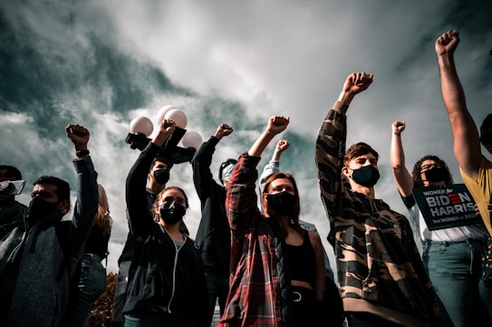 A group of people wearing masks, with some raising their fists in the air. They appear to be at a public gathering or protest. The sky is overcast, adding a dramatic effect to the scene. One person holds a sign supporting a political figure.