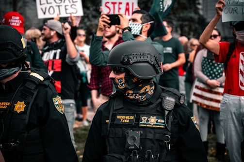 A group of law enforcement officers stands amidst a crowd of protesters. The officers are in tactical gear, including helmets and radios. People in the background hold signs, with some reading 'Legal Votes' and 'Steal', and many wear masks and casual clothing. The setting appears tense with individuals capturing the scene on their mobile phones.