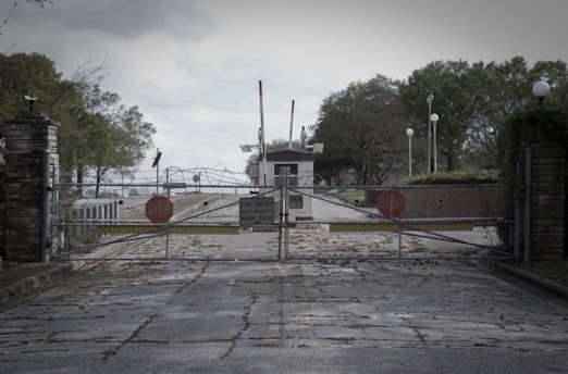 A closed metal gate with several 'stop' signs blocks the entrance to a guarded area. A small guardhouse is visible in the background, with trees and lampposts lining the pathway. The ground is rough and slightly overgrown, suggesting limited maintenance.