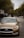 A gold-colored sedan is prominently positioned on a suburban street with trees lining the sides. The background includes other vehicles and residential buildings under a cloudy sky.