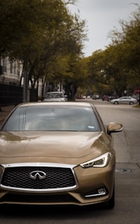 A gold-colored sedan is prominently positioned on a suburban street with trees lining the sides. The background includes other vehicles and residential buildings under a cloudy sky.