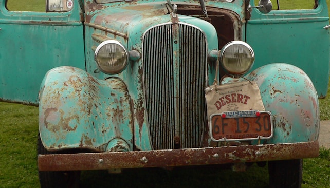 An old, worn-out car with a rusty exterior and faded teal paint. The front of the car features a chrome grille and round headlights. There is a license plate with the number '6F 1530' and a small sign reading 'DESERT' on the bumper. The car is parked on a grassy area with one door open.