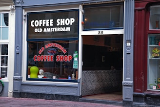 The exterior of an establishment called 'Old Amsterdam Coffee Shop' with large windows displayed with signage. The decor includes a mannequin bust, a green circular object, and zebra stripe pattern seating inside. The door and windows are framed by ornate molding, set between a white and maroon building front.
