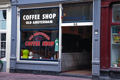 The exterior of an establishment called 'Old Amsterdam Coffee Shop' with large windows displayed with signage. The decor includes a mannequin bust, a green circular object, and zebra stripe pattern seating inside. The door and windows are framed by ornate molding, set between a white and maroon building front.