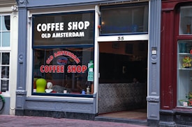 The exterior of an establishment called 'Old Amsterdam Coffee Shop' with large windows displayed with signage. The decor includes a mannequin bust, a green circular object, and zebra stripe pattern seating inside. The door and windows are framed by ornate molding, set between a white and maroon building front.