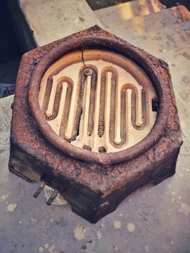 A rusted, hexagonal metal object with a coiled heating element inside is placed on a concrete surface. The object appears weathered, with significant rust and cracks visible in its structure. The coil inside is arranged in a meandering pattern.