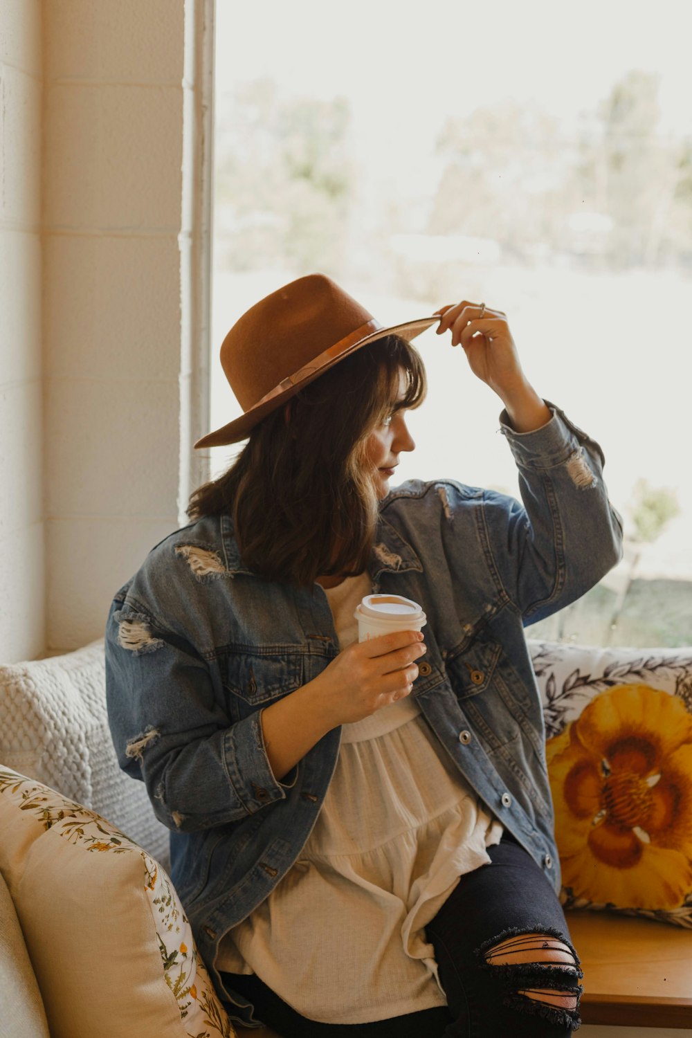 woman in blue denim jacket and brown hat sitting on white and brown floral sofa