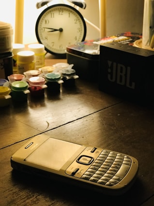 A vintage Nokia phone with a QWERTY keyboard is placed on a wooden table. Nearby, there is an analog clock with large black numbers, multiple small paint containers, and a JBL speaker. The scene is softly lit with warm light, creating shadows on the surface.