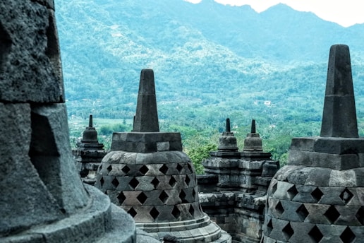 Large stone stupas with intricate patterns are set against a backdrop of lush green hills and mountains, capturing the essence of ancient cultural architecture.