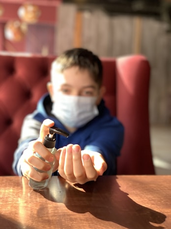 A child wearing a face mask is seated on a red cushioned chair, holding a bottle of hand sanitizer, and applying it to their hands. The background is softly blurred, drawing focus to the action of using the sanitizer, with light illuminating the scene.